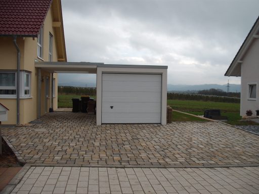 Garagenzufahrt mit Pflastersteinen, angrenzendes Wohnhaus und bewölkter Himmel.