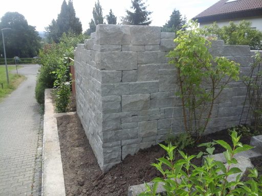 Mauer aus hellgrauen Steinen, umgeben von Pflanzen und einem gepflasterten Weg.