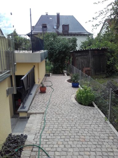 Pflasterweg entlang eines Wohnhauses mit Töpfen und Garten im Hintergrund.